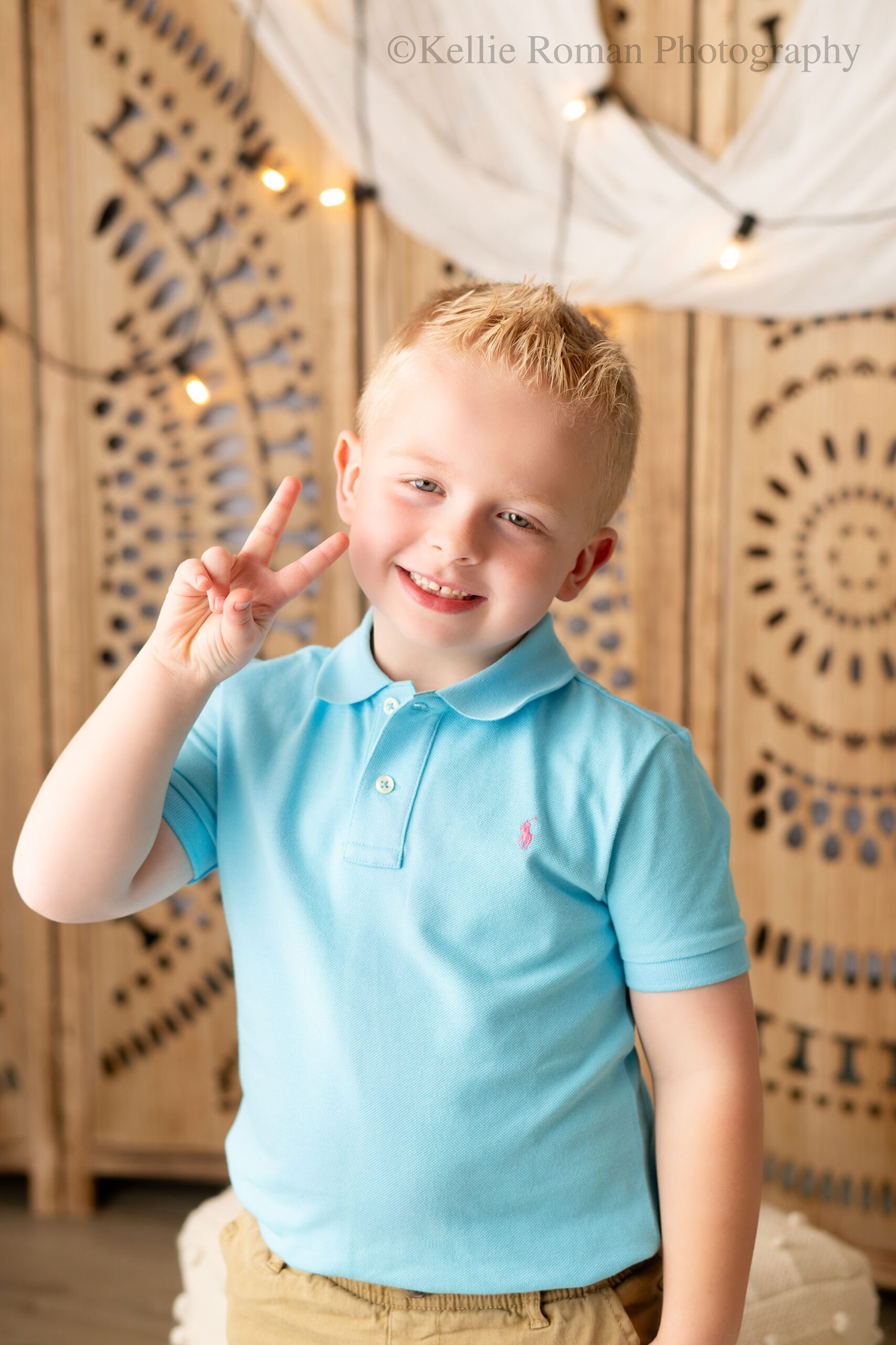 photographer near me. six year old blond boy is standing in front of decorative wood with lights on it. he has a blue polo shirt on and is giving the peace sign. 