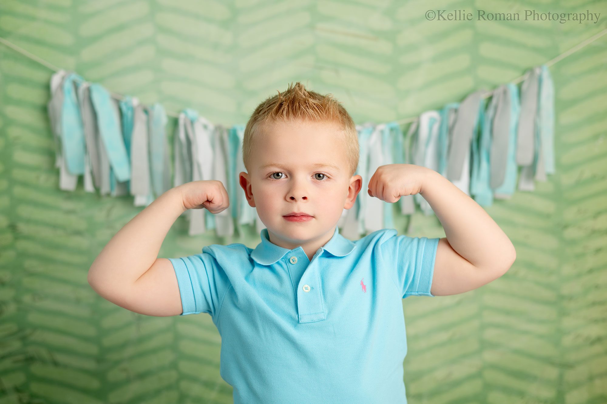 photographer near me. blonde boy is making muscles and has a serious face while looking at camera. he has a light blue polo shirt on. the backdrop is green with a blue and grey fabric banner. 