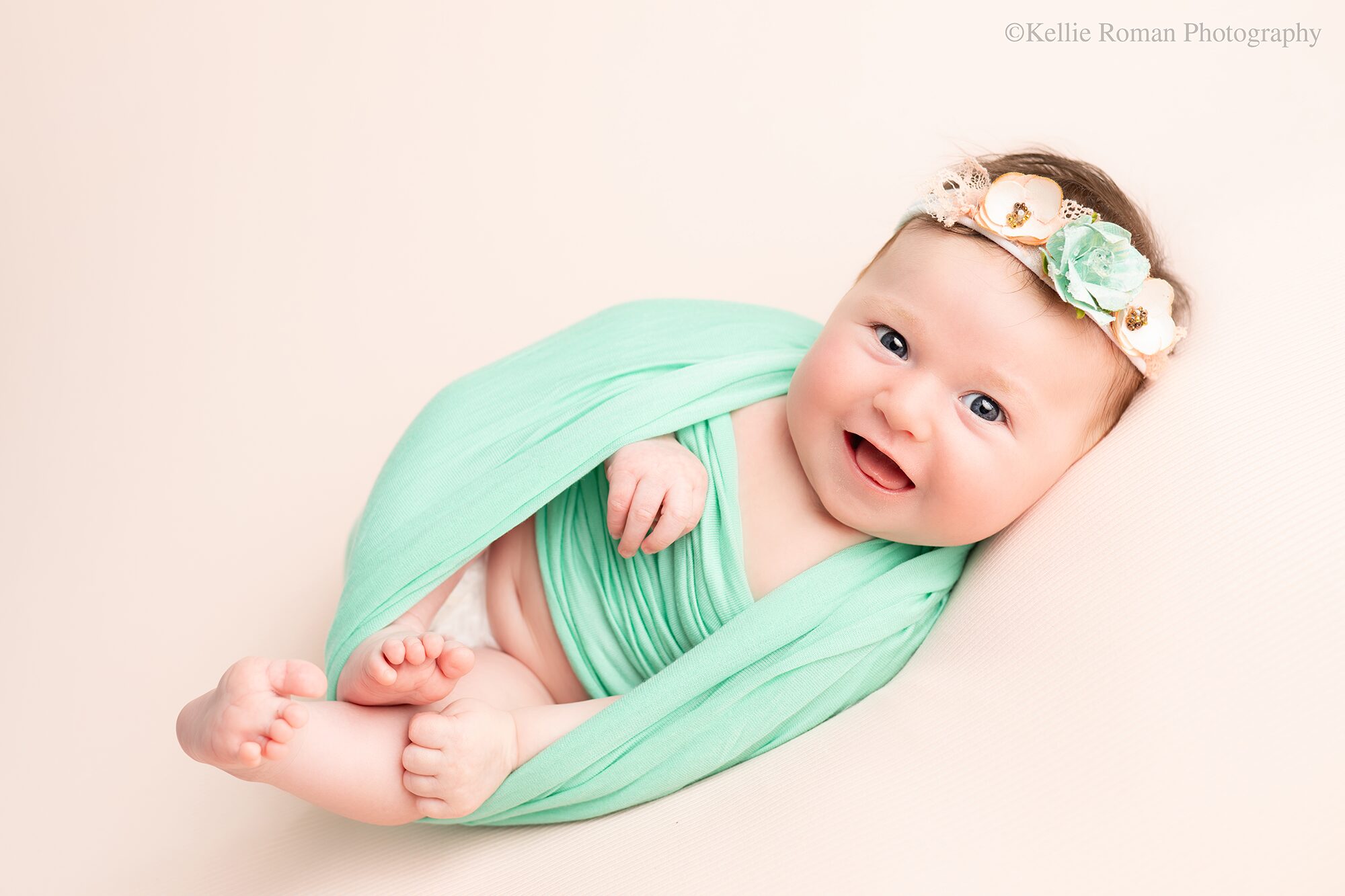 experienced newborn photographer. baby girl wrapped in a teal swaddled is smiling big laying onto of a pink cloth. she has a teal and pink headband on.