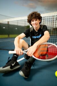 milwaukee senior portraits. a high school senior boy is sitting on a blue tennis court he's holding onto his tennis racket, and wearing his tennis uniform. senior is resting his back against the tennis net and smiling.
