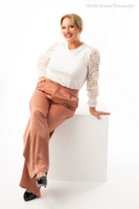 milwaukee headshot photography. women is sitting on white box in front of white backdrop. she has silky blush dress pants on with black heels and white lace top. one hand is in her pocket and she's leaning on the other hand.