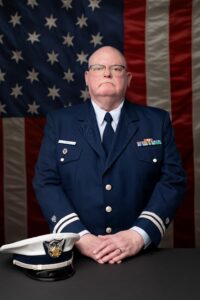 milwaukee headshot photographer. man in coat guard uniform is standing in front of American flag. his hands are resting on grey ledge and his coast guard hat is sitting near his hands.