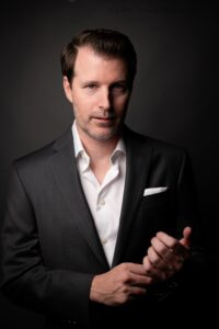 milwaukee headshot photographer. man wearing a dark grey suit with un buttoned white shirt underneath is holding his hands in front of him and smiling at camera. the backdrop is dark grey.