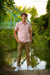 greendale senior photography. high school senior boy is standing in front of a small river with a teal bridge behind him and lots of green leaves. he has his hands in his pockets of his khaki pants and is wearing a pink button up shirt.