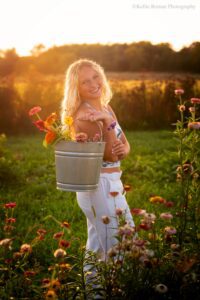 oak creek senior pics. high school girl is standing in flower field holding a metal bucket filled with flowers over her shoulder. the sun Is setting behind her. she has long blonde hair, and is wearing white pants with a white and blue tank top.