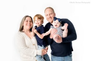 milwaukee family pictures. family of four is standing in milwaukee photo studio. mom is holding young son and dad is holding their baby daughter. family has jeans on and shades of navy and cream. they are all smiling at the camera. baby girl has her hands and feet sticking out, and is holding onto one of her feet.