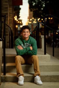 oak creek senior photographer. milwaukee senior guy is sitting on stairs in downtown milwaukee. he is crossing his arms while resting them on his knees. he has khaki pants on with a green sweater. there are lights behind him for a milwaukee restraunt.