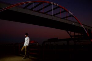 milwaukee senior pictures. high school senior guy leaning against red railing with his hands in his pockets. he's standing underneath milwaukee hoan bridge at night. he's wearing white button up shirt and khakis. the bridge is also lit up.