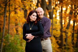 Milwaukee Family Photographer. a married couple is standing in milwaukee park in the fall. the backdrop is yellow and green leaves. the husband has his arms around his wife. women is wearing a black dress, the man has a plaid shirt on.