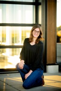 milwaukee senior photographers. high school senior from New Berlin is kneeling on concrete in front of glass building. the sun is setting and shining through the building. girl has jeans on with a black top and wire framed glasses.