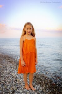 milwaukee family photographer. young girl standing on rocky shore. Lake Michigan is behind her. she has an orange dress on.