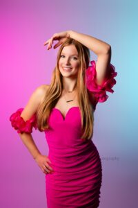 milwaukee premium senior photography. high school girl with hot pink dress is standing with one hand on hip and the other over her head. she has long hair and the backdrop is pink and blue colors.