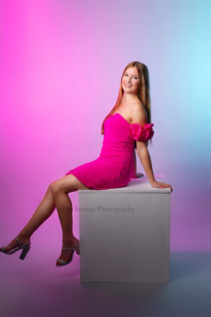 milwaukee premium senior photography. high school girl wearing bright pink dress is sitting on white cube with her hands on it, behind her. she has silver heels on and is dangling her legs. she has long hair, and is smiling. the backdrop are fun colors and lights of pink and blue.