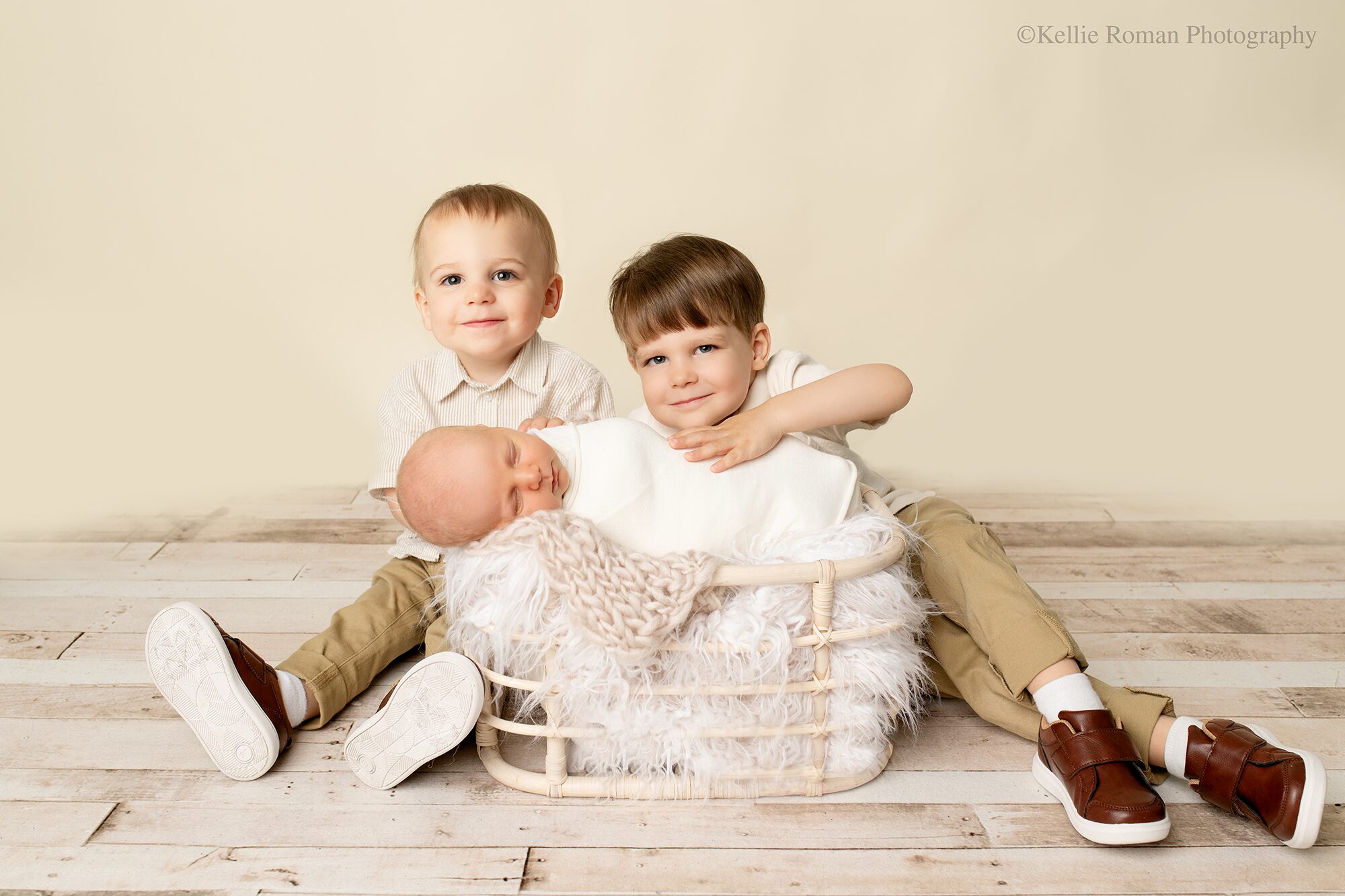 Waukesha family photographer. three brothers are sitting on wood floor with their newborn brother in a wood basket stuffed with fur. older boys are sitting behind basket. they are looking at the camera and smiling, while baby brother is sleeping.