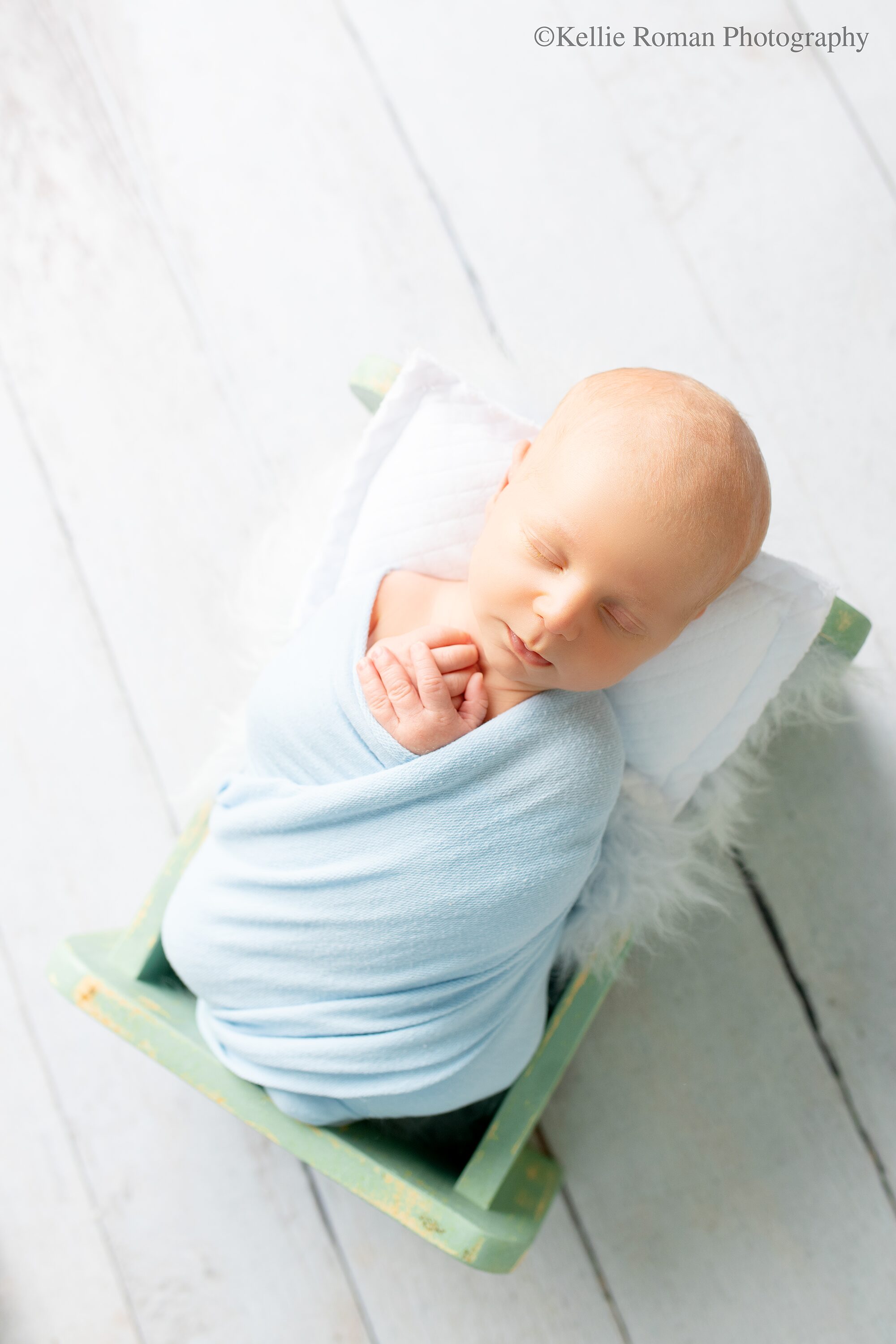 waukesha newborn photographer. newborn boy is asleep and wrapped in a light blue swaddle. his hands are sticking out and resting on his chest. he's laying on his back in a teal bed with a white pillow under his head.