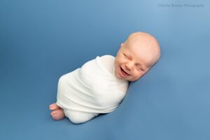 Waukesha newborn photographer. newborn boy in a white swaddle is laying on his side ontop of a blue fabric. his feet are sticking out and he is smiling very big.