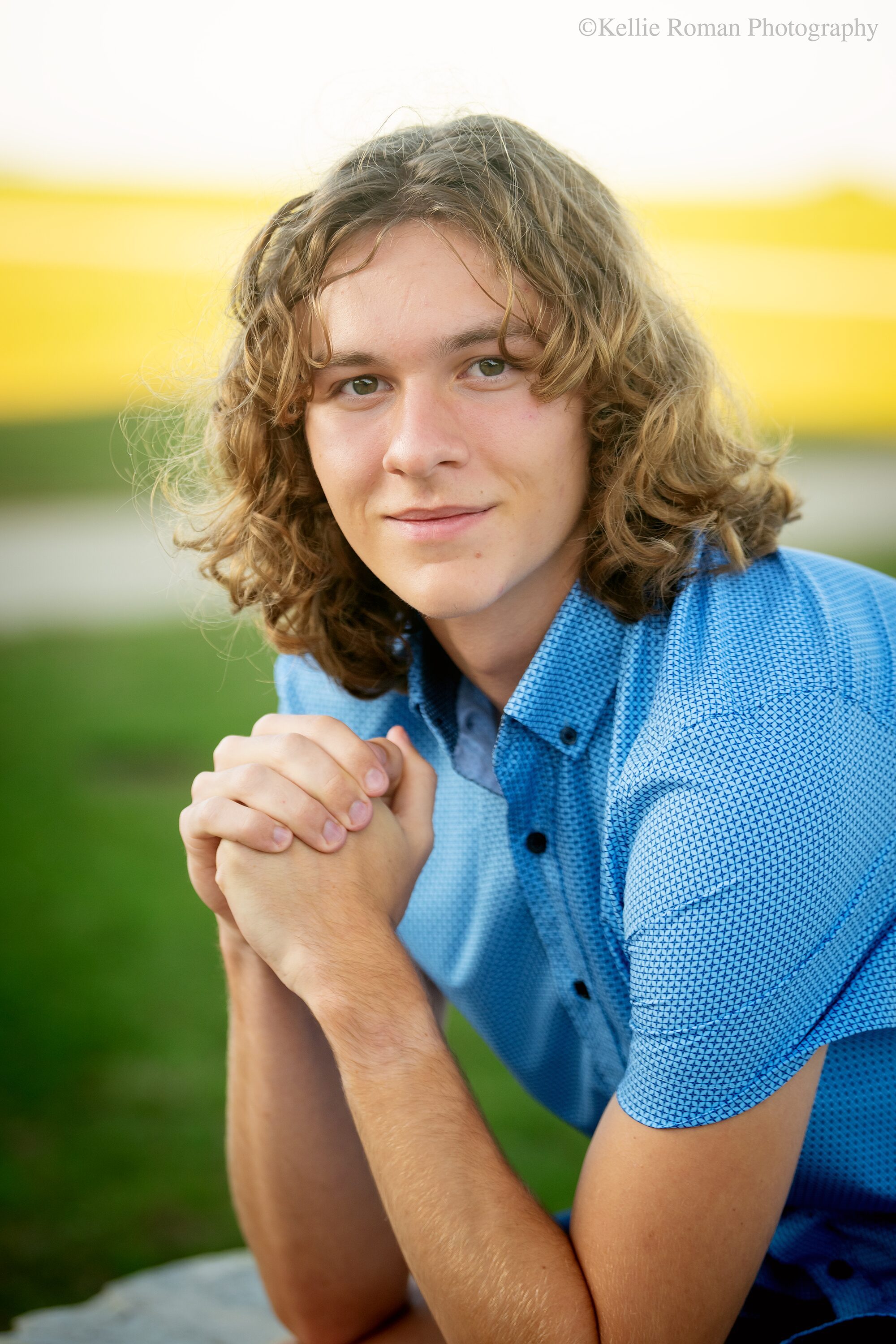 milwaukee family and senior photographer. high school senior guy is in milwaukee park. he has his hands up and folded while smiling at the camera. he has a blue shirt on and curly hair.