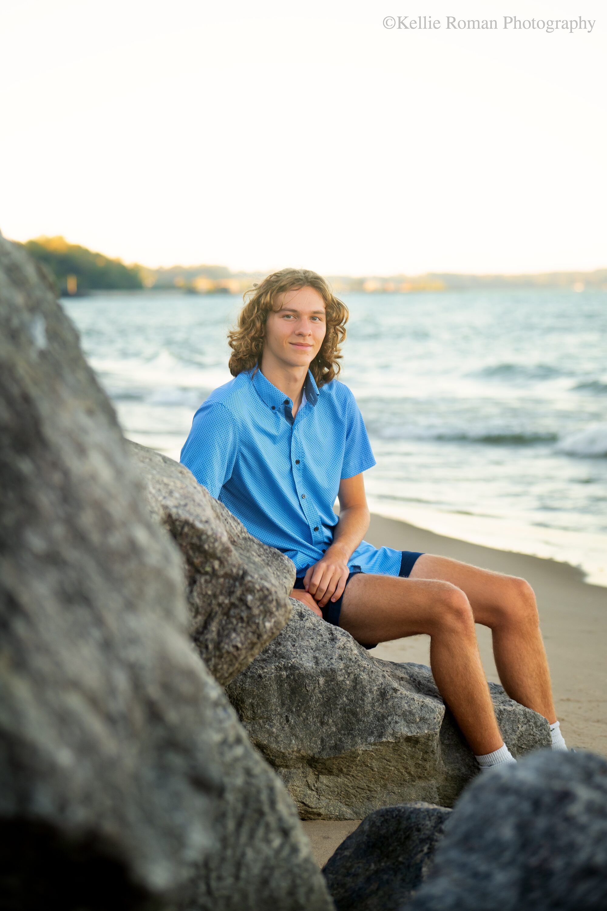 milwaukee family and senior photographer. high school senior guy is sitting on rocks on beach with Lake Michigan behind him.