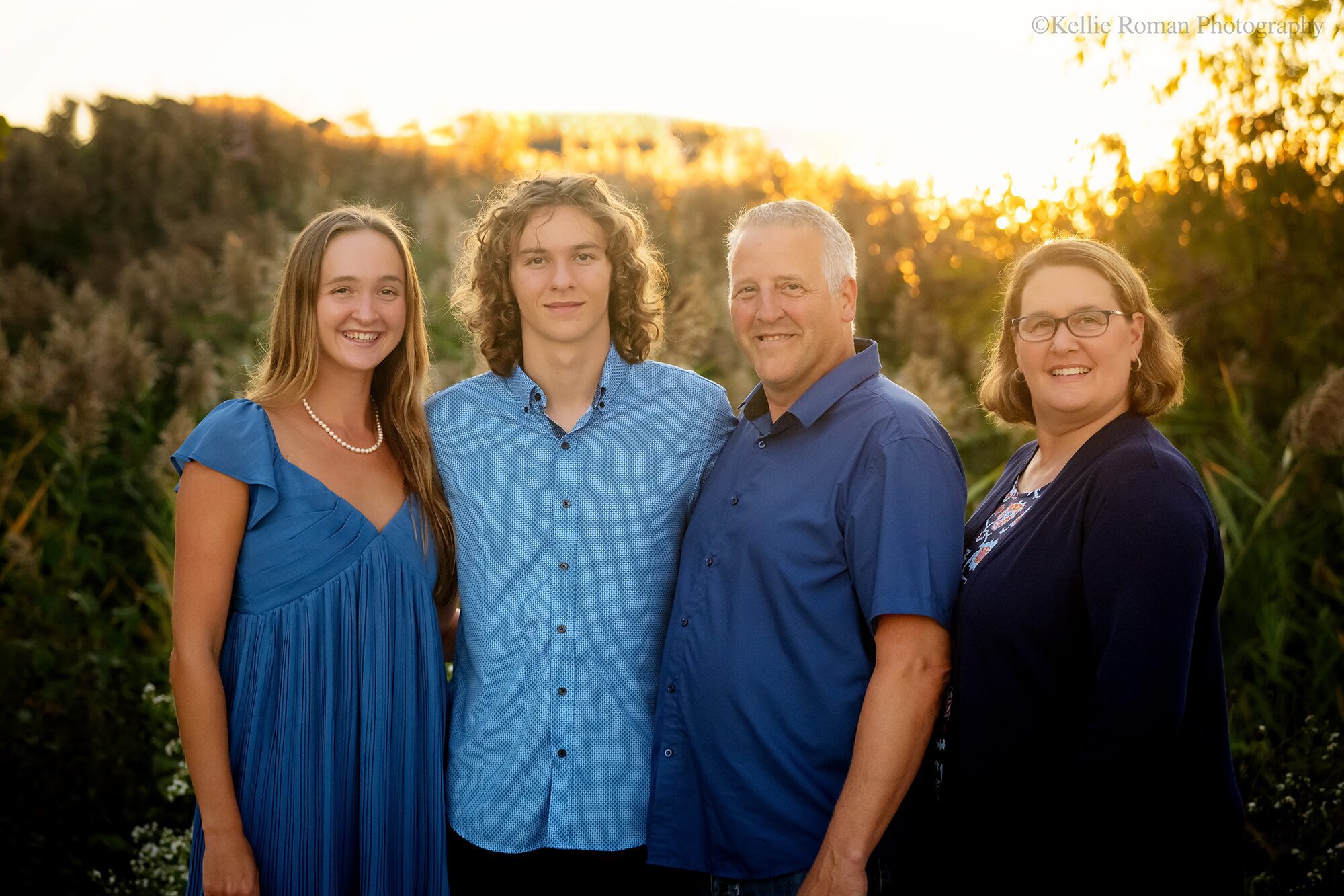 milwaukee family and senior photographer. family of four is standing in park in front of tall grass with the sun setting behind them. they have shades of blue on.