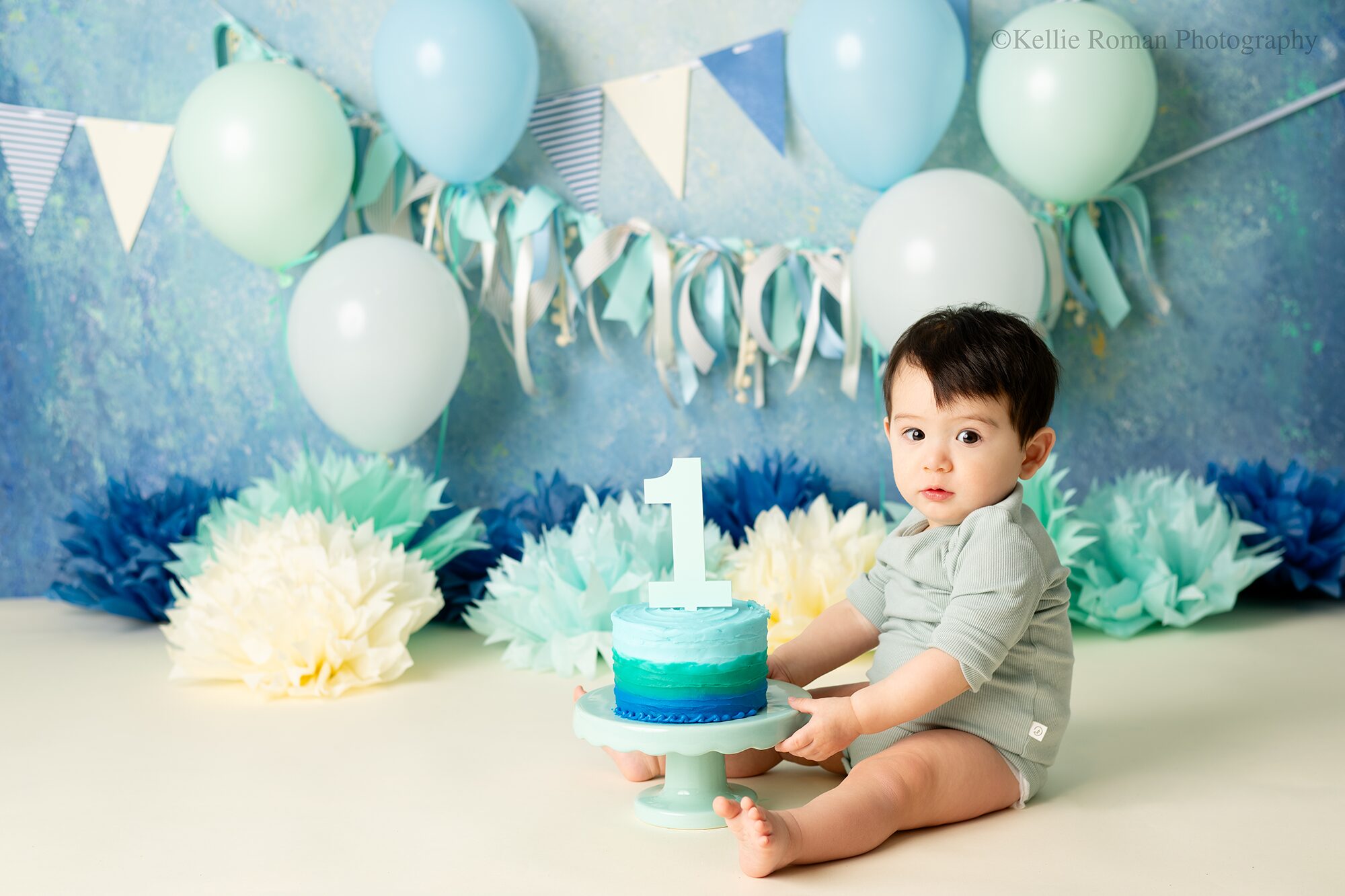 colorful milwaukee cake smash. one year old boy with blue romper on is sitting on floor with hands on teal cake stand. teal and green ombre cake is on stand. backdrop has balloons banners and tissue paper on the floor in blues and creams.