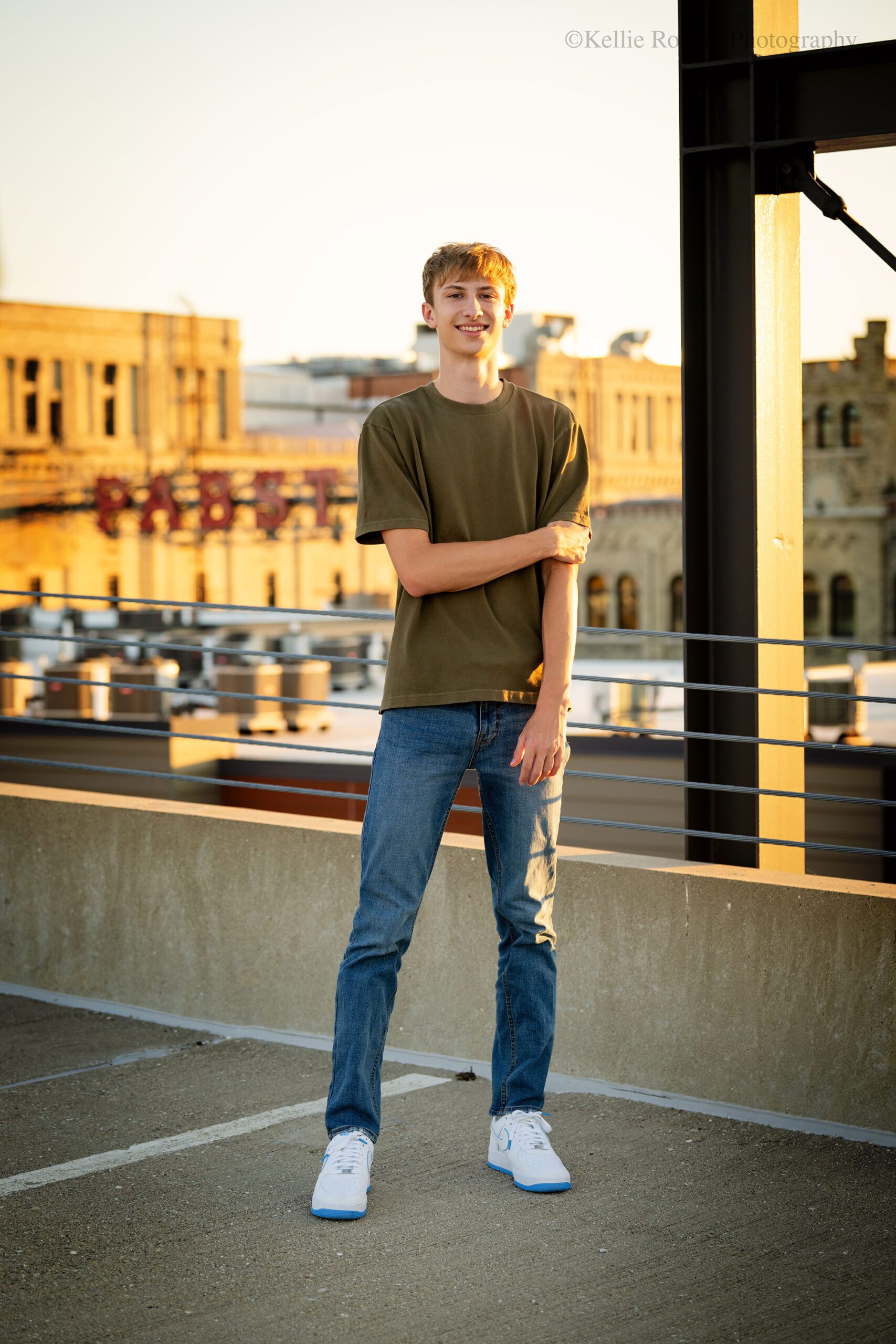 milwaukee senior photographers. oak creek senior standing on parking garage rooftop in downtown milwaukee. the Pabst sign is behind him and a large metal structure. he has on jeans and a t shirt. 