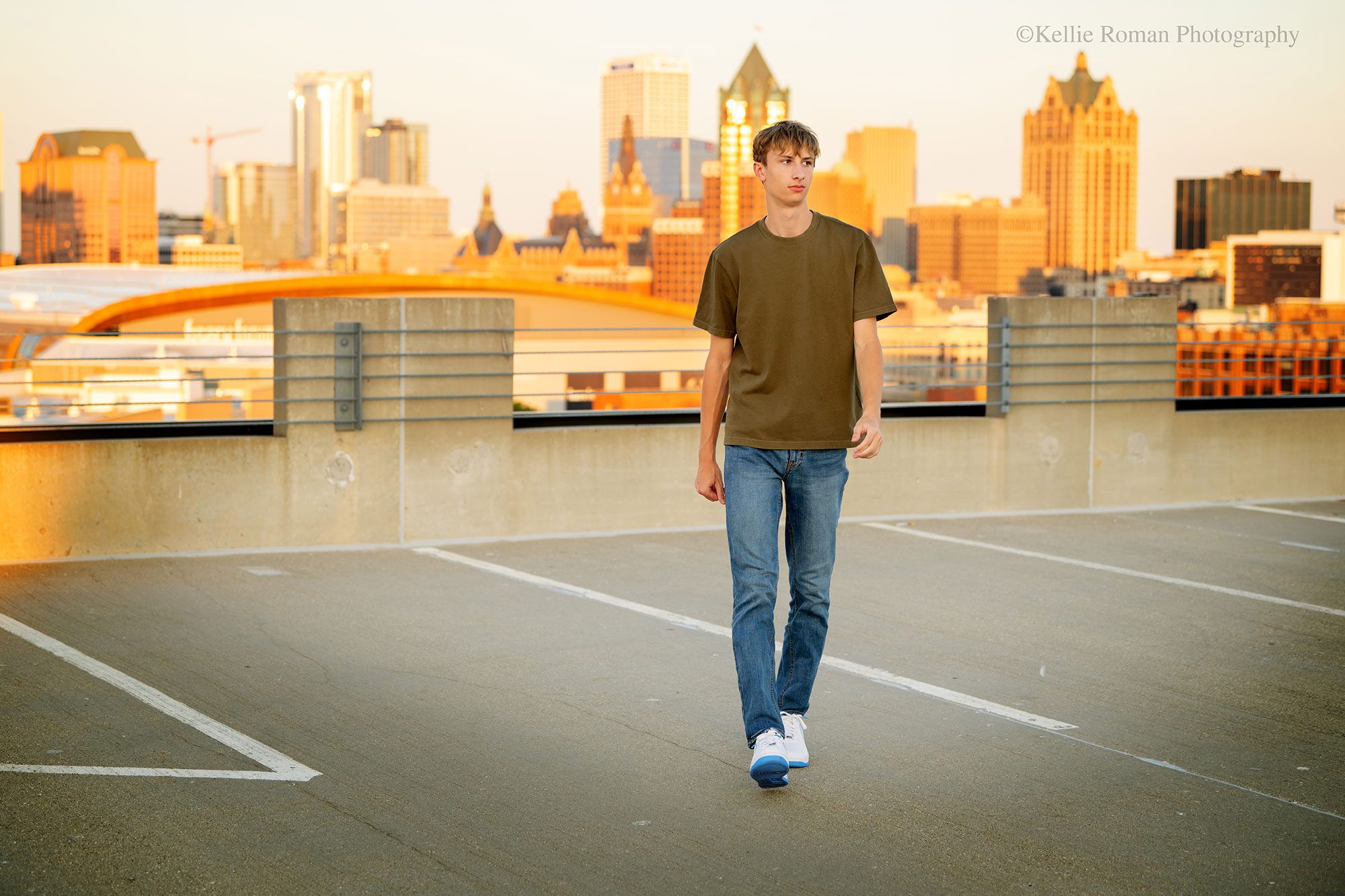 best milwaukee senior photos. oak creek senior walking and looking to the side on top of a parking garage in downtown milwaukee. the milwaukee skyline is behind him in the distance and it's light up orange because of the setting sun. 