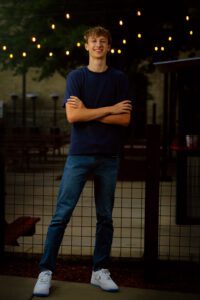 milwaukee senior photographers. oak creek senior guy is standing in his arms crossed and leg out in front of patio lights behind him.