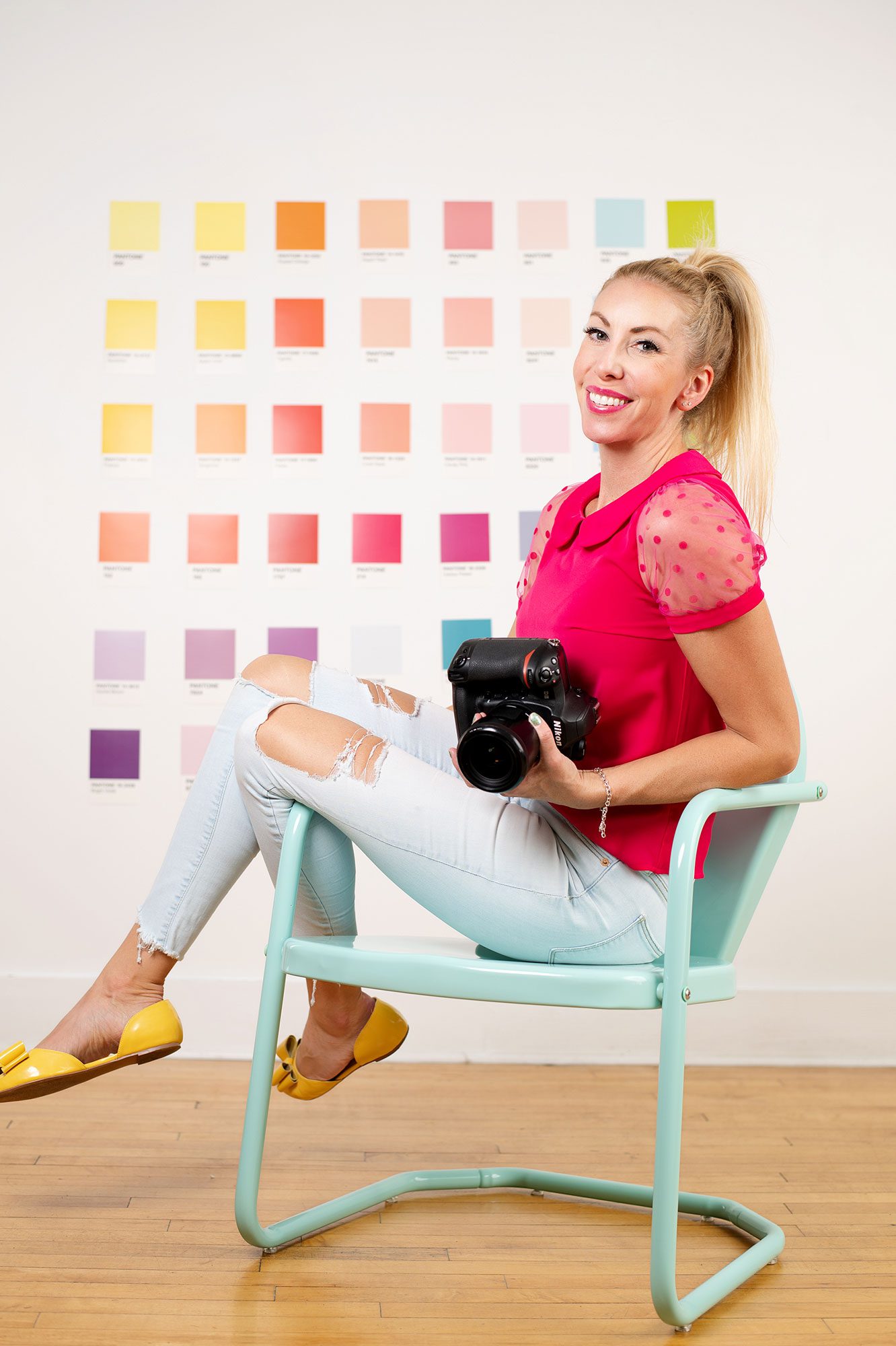 Kellie Roman Photography. Milwaukees best photographer. Kellie is sitting on a teal chair holding her camera with bright colors behind her. she has a pink top on with jeans and yellow shoes.