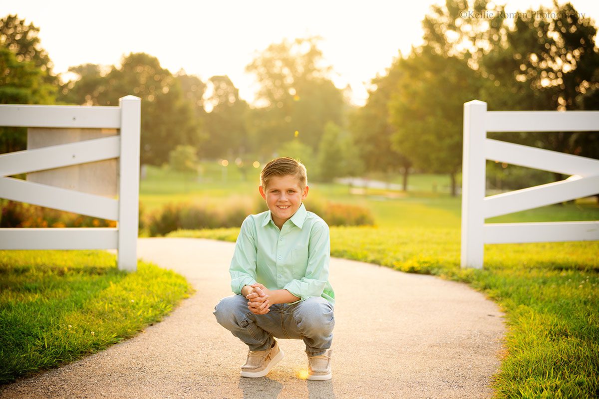 Birthday photo session. ten year old boy is squatting on path where white picket fence is behind him with the setting sun.
