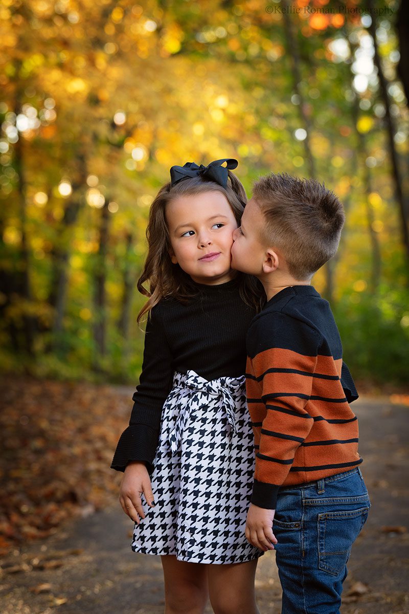 milwaukee family photographer. a brother and sister are standing in a park and brother is kissing sister on the cheek. the girl is looking off to the side. 