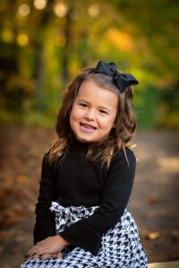 milwaukee family photographer. young girl sitting on wood bench in milwaukee park. she has a black and white dress on and has her hands in her lap. the glowing sun is behind her through the fall colored trees.