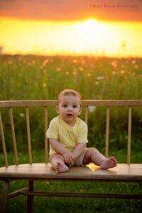milwaukee family photographer. baby boy sitting on wood bench in milwaukee park. he's has a yellow shirt on and shorts. the sun is setting behind him and is glowing orange.