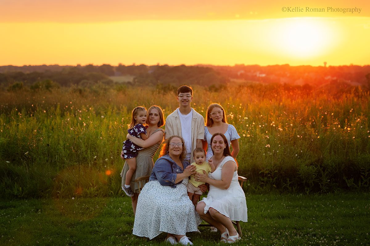 milwaukee family photographer. sky has bright orange sunset with multiple family members and young children sitting and standing while looking at camera.