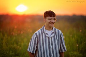 milwaukee family photographer. high school boy standing in park with glowing orange sunset behind him. he's wearing a striped shirt and glasses and is looking off to the side.