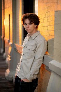 oak creek senior pics. high school senior guy leaning against white cream brick building in downtown milwaukee. golden orange sun is shining onto the brick. he has his hands in his jean pockets.